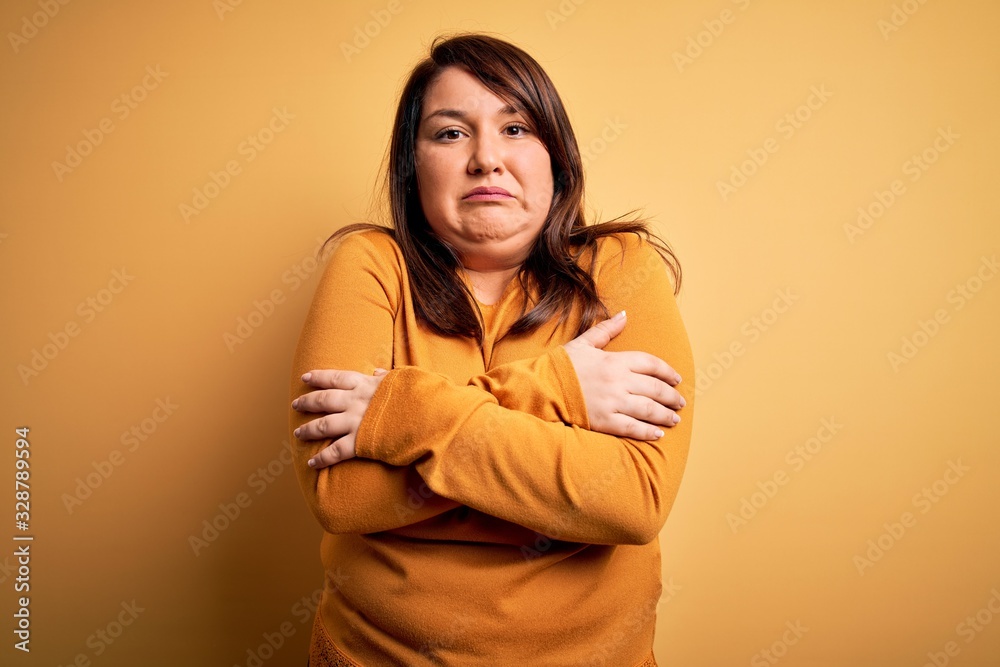 Beautiful brunette plus size woman wearing casual sweater over isolated yellow background shaking and freezing for winter cold with sad and shock expression on face Beautiful brunette plus size woman wearing casual sweater over isolated yellow background shaking and freezing for winter cold with sad and shock expression on face