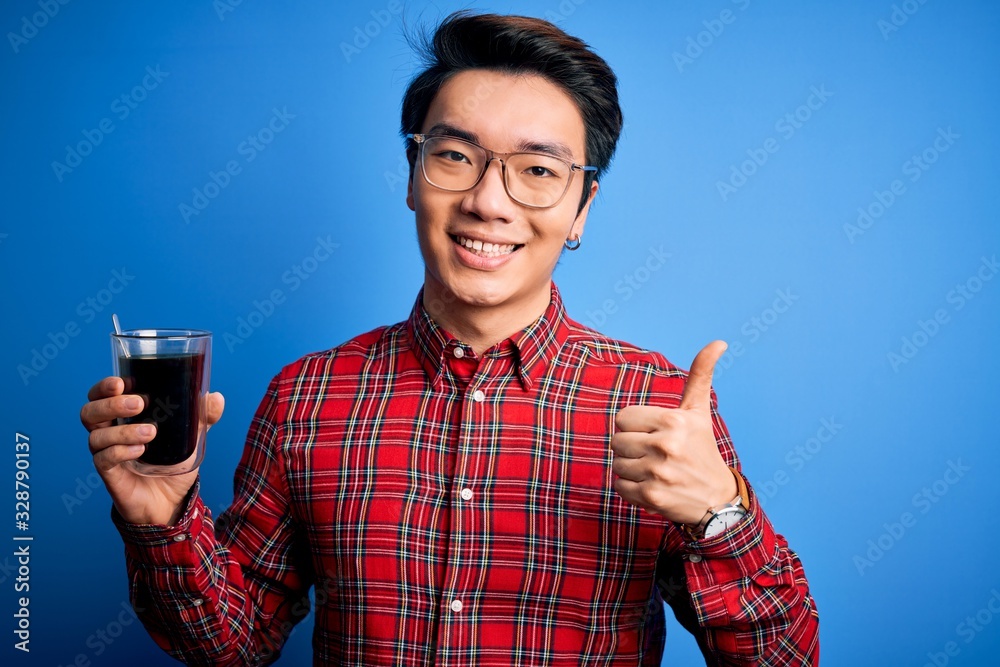Young handsome chinese man drinking glass of coffee over isolated blue background happy with big smile doing ok sign, thumb up with fingers, excellent sign Young handsome chinese man drinking glass of coffee over isolated blue background happy with big smile doing ok sign, thumb up with fingers, excellent sign