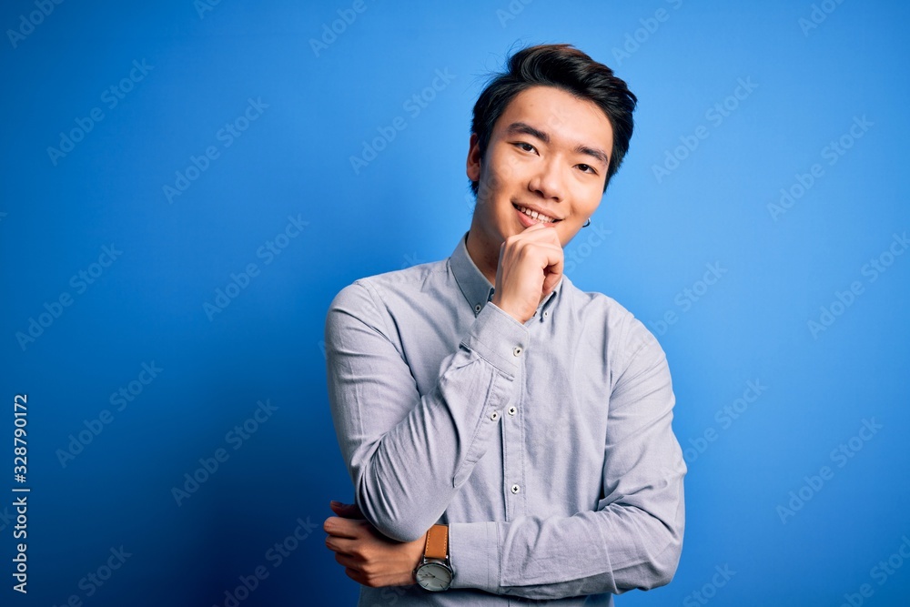 Young handsome chinese man wearing casual shirt standing over isolated blue background looking confident at the camera smiling with crossed arms and hand raised on chin. Thinking positive. Young handsome chinese man wearing casual shirt standing over isolated blue background looking confident at the camera smiling with crossed arms and hand raised on chin. Thinking positive.