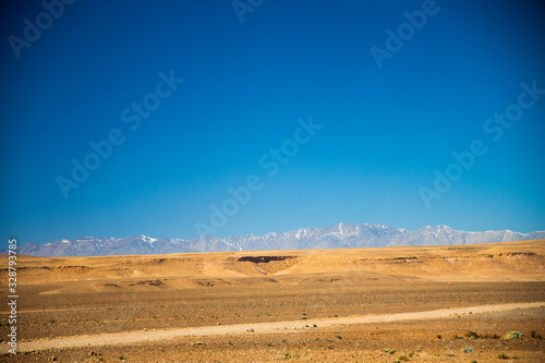 Desert road lies among the valley with Atlas mountains and blue sky background