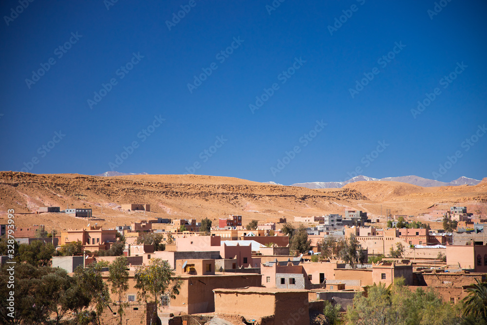 Berber village full of trees and sand lies at bottom of the desert hill with the mountains background