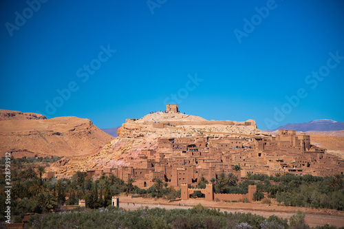 Ancient kasbah full of people lays between green oasis and hills with mountains background