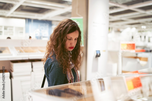 Young caucasian woman with curly black hair at the shopping mall store looking products price on the shelf standing alone