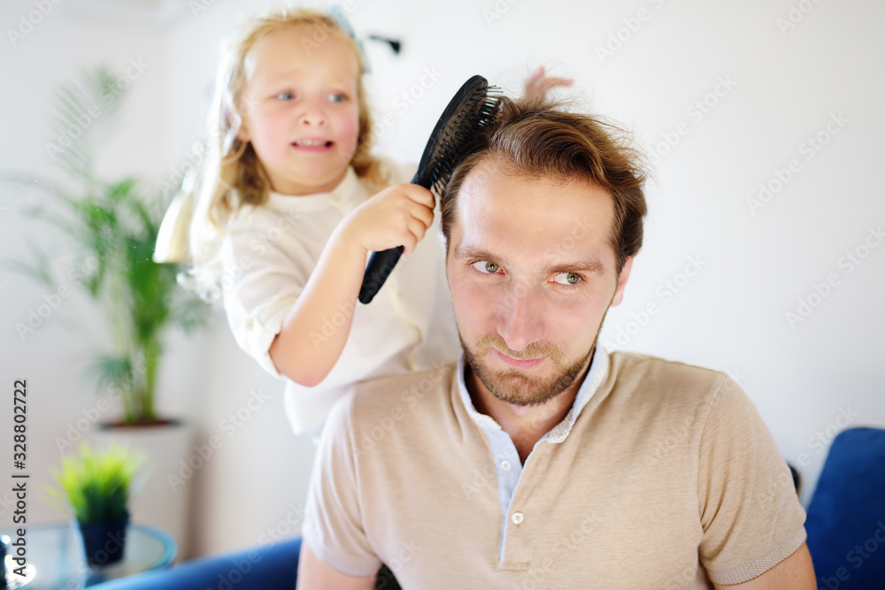 Little curly hair daughter making funny hairstyle for her young handsome father. Preschooler girl brush hair of her dad and play as if she works in a beauty salon Little curly hair daughter making funny hairstyle for her young handsome father. Preschooler girl brush hair of her dad and play as if she works in a beauty salon