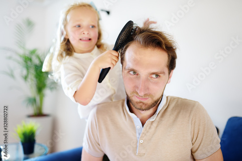Little curly hair daughter making funny hairstyle for her young handsome father. Preschooler girl brush hair of her dad and play as if she works in a beauty salon