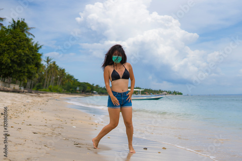 use of medical face mask in public places- young attractive Asian Chinese woman enjoying beach holidays in bikini and protective facial mask in prevention vs virus infection
