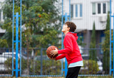 Cute boy in red t shirt plays basketball on city playground. Active teen enjoying outdoor game with orange ball. Hobby, active lifestyle, sport for kids.
