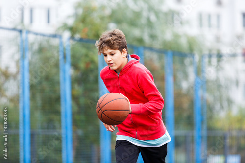 Cute boy in red t shirt plays basketball on city playground. Active teen enjoying outdoor game with orange ball. Hobby, active lifestyle, sport for kids.