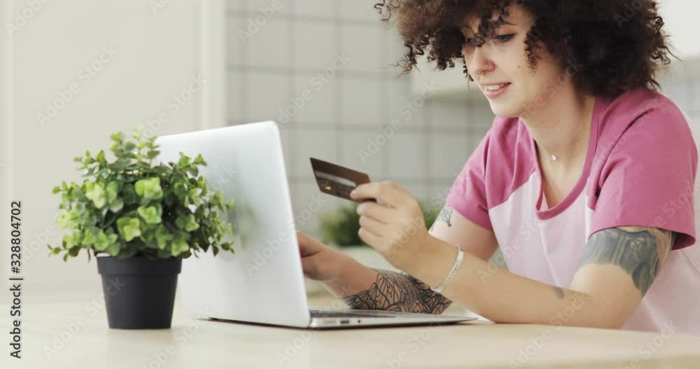 Happy woman is making online payment holding bank card using modern laptop at home. Finance, online shopping in internet concept. Curly hairs brunette girl is making online purchase.