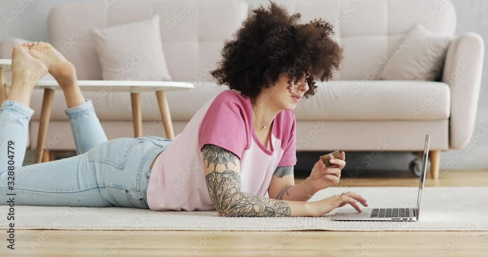 Young woman making online payment holding bank card using modern laptop at home lying on floor in living room. Finance, online shopping concept. Girl with brunette curly hairs doing online purchase. Young woman making online payment holding bank card using modern laptop at home lying on floor in living room. Finance, online shopping concept. Girl with brunette curly hairs doing online purchase.