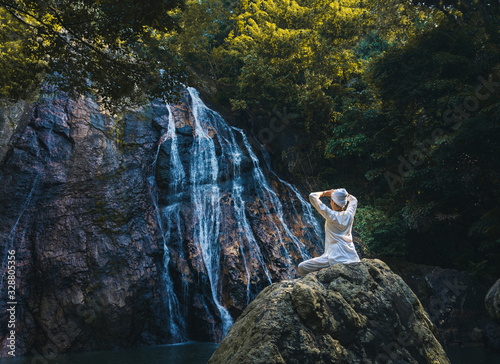 Young meditating woman in a white clothes and turban sitting on the rock by the waterfall in the forest