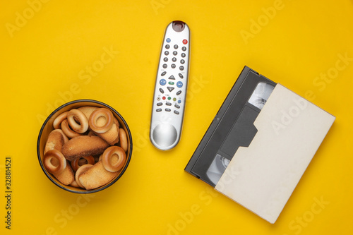 TV remote control and bowl of crackers, video cassette on yellow background. Watching the TV. The concept of home rest after work. 80s. Top view. photo