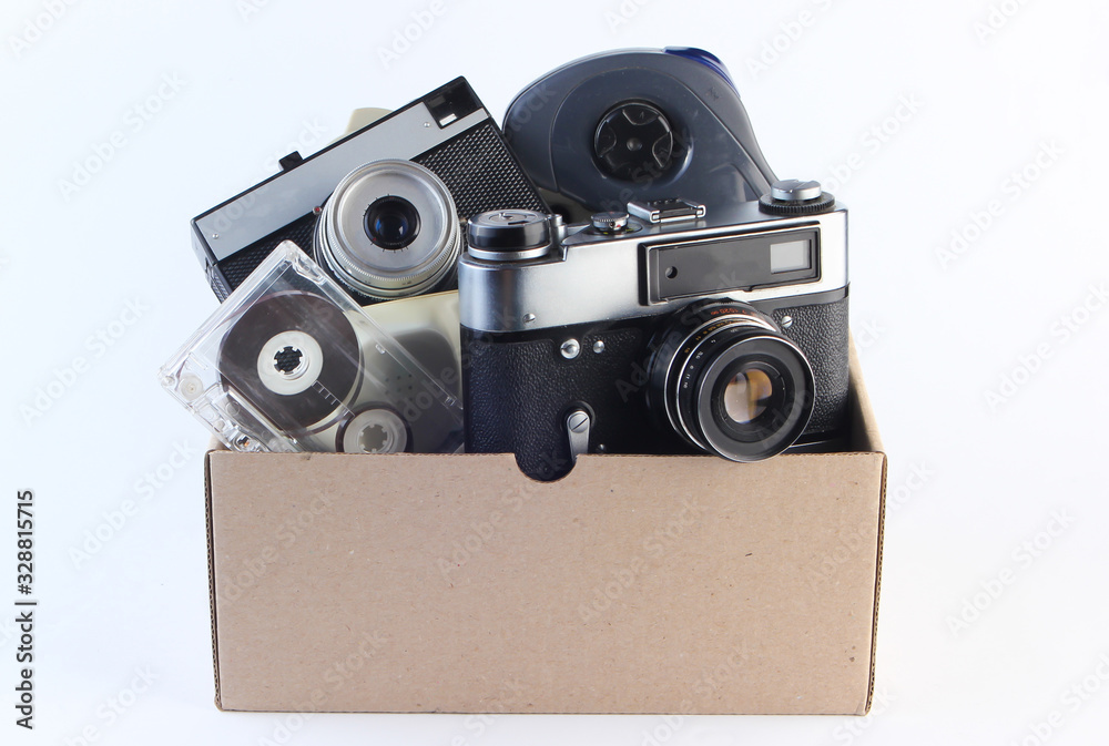 Cardboard box with retro film camera, audio cassette, gamepad on a white background. Antiques, trash