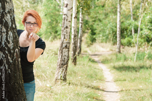 Shadowing and surveillance concept with copy space. Beautiful ginger girl stand behind tree and point finger to camera. Young woman in summer birch forest.