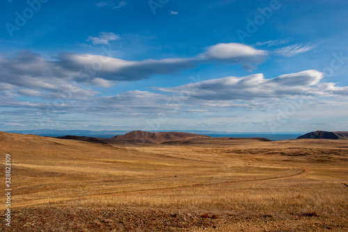 Steppe with hills, clouds in the sky and mountains in the distance. Country road. Brown and yellow grass. Blue sky with fluffy clouds.