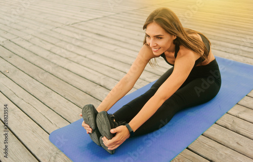 Young fitness woman doing stretching holding legs while sitting on a yoga mat on wooden boards outdoors © splitov27