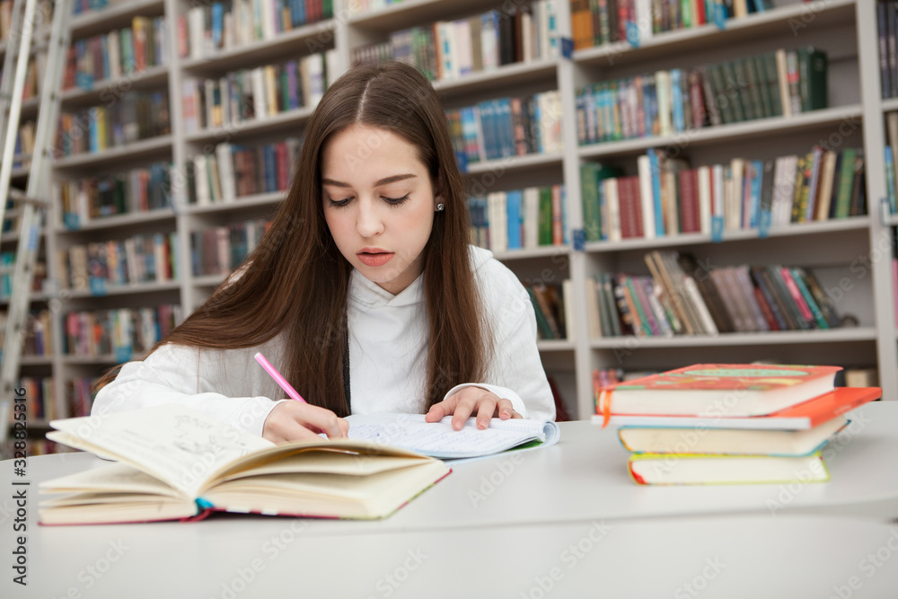 Young teenage girl looking concentrated, writing in her textbook at the library, Female teen student working on assignment for school Young teenage girl looking concentrated, writing in her textbook at the library, Female teen student working on assignment for school