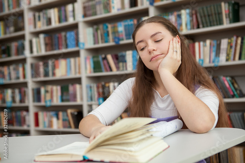 Teenage girl looking tired or bored, studying at the library. Exhausted teen student preparing for exams, reading a book