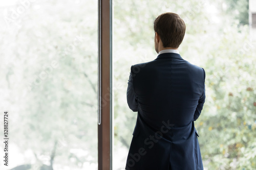 Back view of confident young businessman in formal suit stand near window look in distance thinking planning, concentrated male boss lost in thoughts, consider opportunities, business vision concept