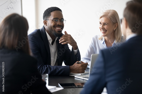 Smiling multiracial diverse businesspeople sit at office desk discussing business project together, happy multiethnic colleagues coworkers brainstorm talk consider cooperation in boardroom