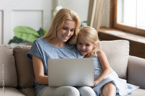 Young Caucasian mom sit on couch relax with little preschooler daughter watch funny videos on laptop together, smiling mother or nanny rest with small girl child using modern computer gadget at home