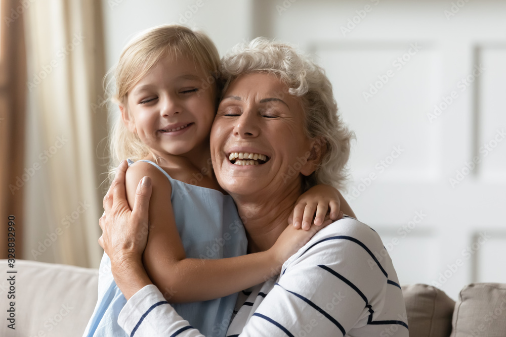 Overjoyed senior grandmother and cute little granddaughter hug cuddle showing love and care, happy mature granny and small girl child embrace enjoy family weekend at home together, bonding concept