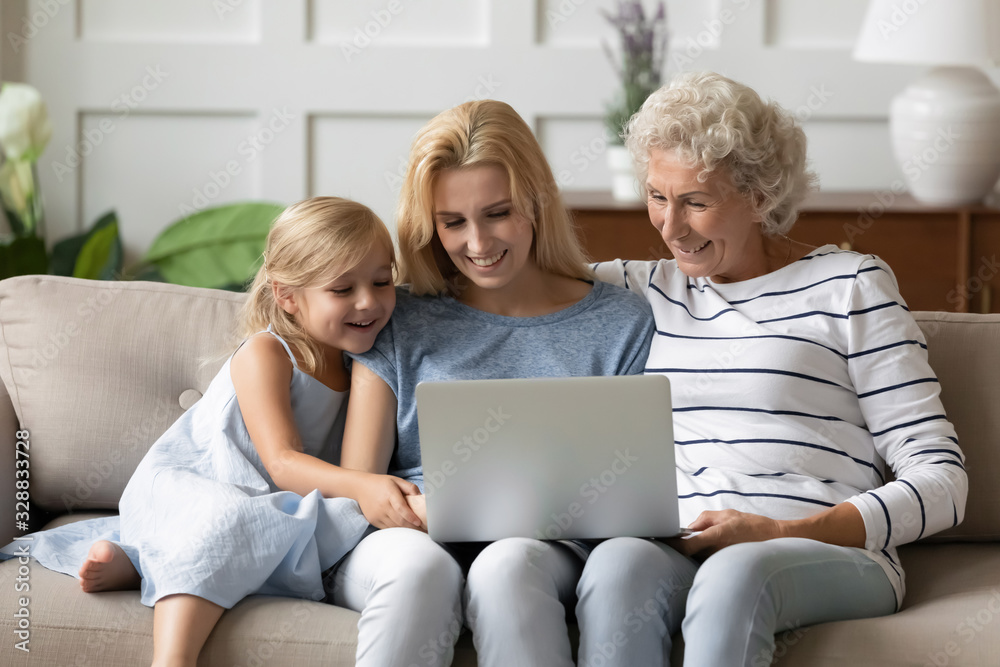 Happy three generations of women sit relax on couch in living room using modern laptop together, smiling little girl with young man and grandmother rest on sofa watching video on computer at home Happy three generations of women sit relax on couch in living room using modern laptop together, smiling little girl with young man and grandmother rest on sofa watching video on computer at home