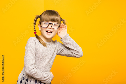 Little child girl journalist in glasses with surprised euphoria face and funny pigtails listening to something holding his hand to ear