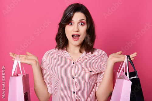 Portrait of young astonished lady standing with shopping bags in hands and amazedly looking directly at camera while posing isolated over pink background, dresses striped blouse, having dark hair. photo