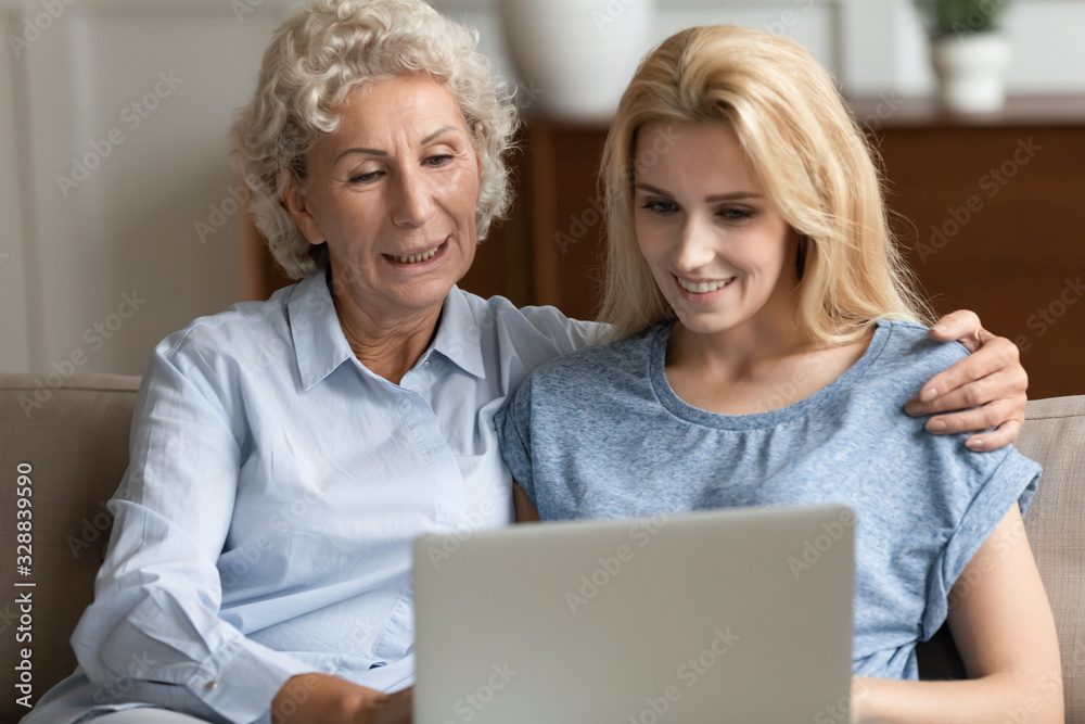 Smiling senior 60s mother and millennial girl child relax at home using modern laptop gadget together, happy mature mom and adult daughter rest on couch watch video browse internet on computer Smiling senior 60s mother and millennial girl child relax at home using modern laptop gadget together, happy mature mom and adult daughter rest on couch watch video browse internet on computer