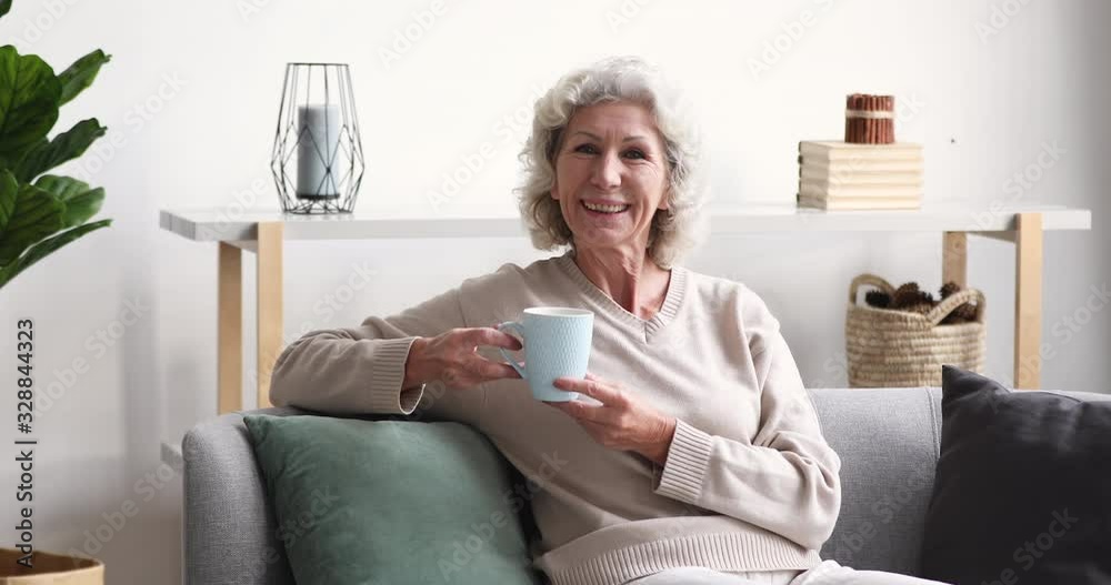Smiling senior adult lady drinking tea looking away at camera at home. Cheerful retired old grey-haired woman laughing relaxing sitting alone on sofa in cozy living room. Happy elder grandma portrait Smiling senior adult lady drinking tea looking away at camera at home. Cheerful retired old grey-haired woman laughing relaxing sitting alone on sofa in cozy living room. Happy elder grandma portrait