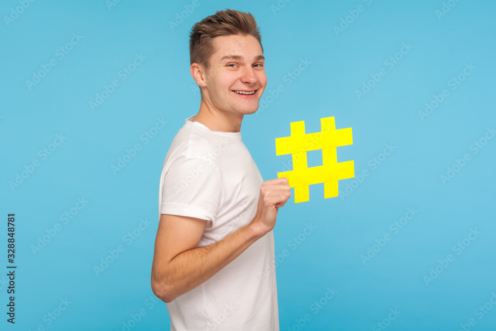 Internet trends. Profile of happy funny man in t-shirt holding big yellow hashtag symbol and smiling, showing hash sign of viral social media topic. indoor studio shot isolated on blue background Internet trends. Profile of happy funny man in t-shirt holding big yellow hashtag symbol and smiling, showing hash sign of viral social media topic. indoor studio shot isolated on blue background