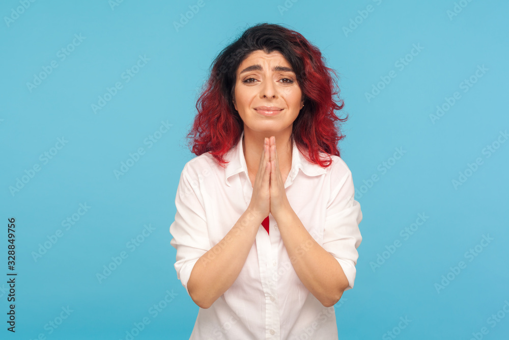 Portrait of depressed unhappy woman with fancy red hair holding hands in prayer and looking imploringly at camera, feeling desperate, asking forgiveness. indoor studio shot isolated on blue background Portrait of depressed unhappy woman with fancy red hair holding hands in prayer and looking imploringly at camera, feeling desperate, asking forgiveness. indoor studio shot isolated on blue background