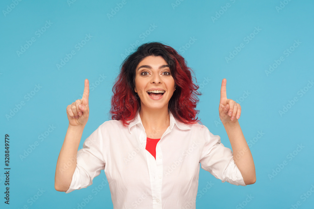 Wow, look up! Astonished hipster woman with fancy red hair pointing above head with amazed shocked expression, showing copy space for commercial text. indoor studio shot isolated on blue background Wow, look up! Astonished hipster woman with fancy red hair pointing above head with amazed shocked expression, showing copy space for commercial text. indoor studio shot isolated on blue background