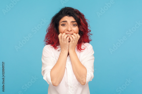 Portrait of scared panicking woman with fancy red hair biting nails and looking with frightened anxious expression, nervous about problems, troubles. indoor studio shot isolated on blue background