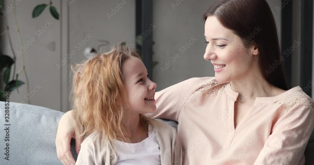 Affectionate young mother and small daughter bonding looking at camera. Loving happy mom hugs cute school child girl sit on sofa. Sweet tender single parent and kid relationship, family portrait