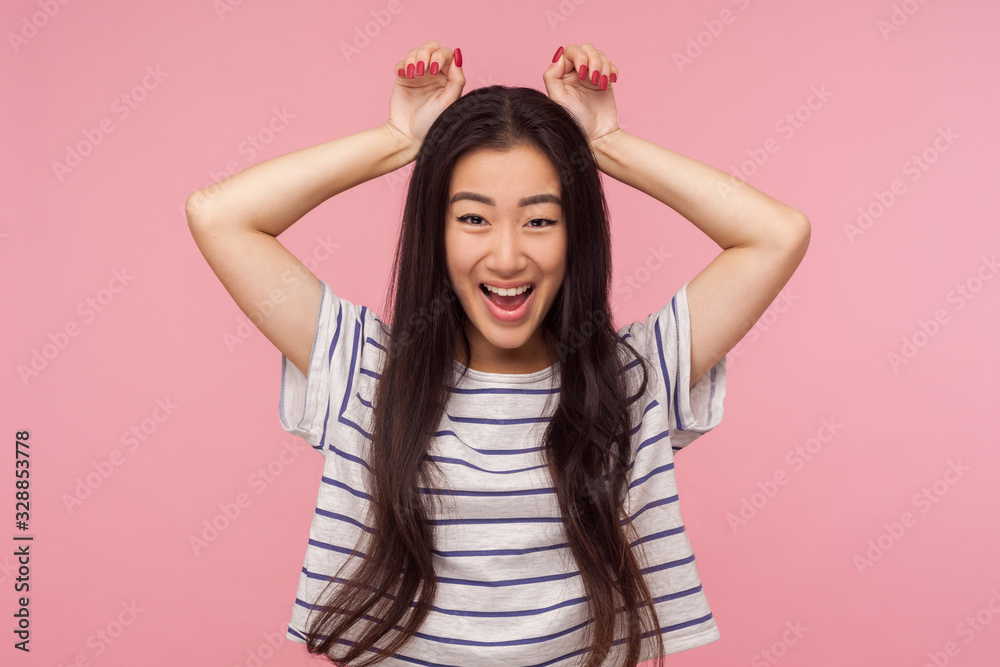 Portrait of childish funny girl with long brunette hair in striped t-shirt showing bunny ears gesture and smiling with playful carefree expression. indoor studio shot isolated on pink background Portrait of childish funny girl with long brunette hair in striped t-shirt showing bunny ears gesture and smiling with playful carefree expression. indoor studio shot isolated on pink background