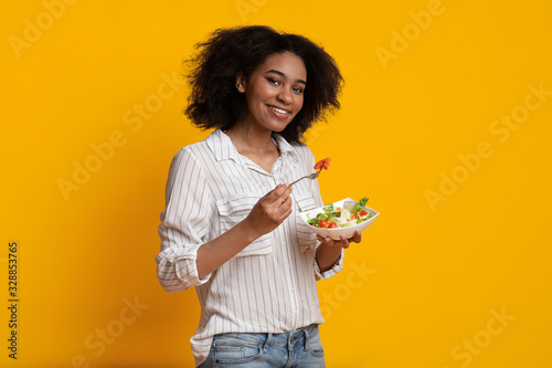 Quadro su tela Smiling Afro Woman Eating Fresh Vegetable Salad Standing On Yellow Background
