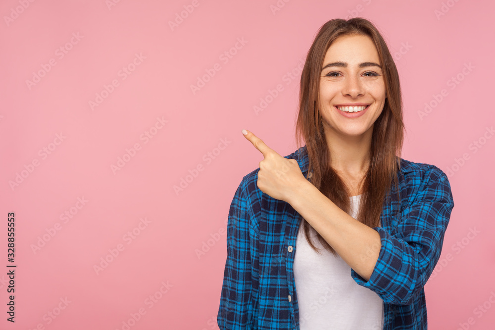 Look at advertisement! Portrait of charming brunette girl in checkered shirt pointing aside at empty place for ad content. studio shot isolated on pink background, copy space for commercial product Look at advertisement! Portrait of charming brunette girl in checkered shirt pointing aside at empty place for ad content. studio shot isolated on pink background, copy space for commercial product