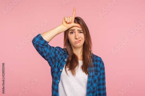 Portrait of depressed unlucky girl in checkered shirt showing loser gesture with fingers on forehead, upset about failure, dismissal at work, blaming herself for mistake. indoor studio shot, isolated photo