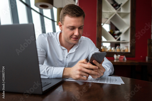 Young businessman wearing a white fashion shirt chatting and talking business on a mobile cell phone in a cafe with a laptop and documents. Freelance and selfemployment concept. Distance job.