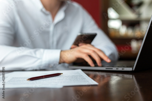Closeup of hands of young businessman wearing a white fashion shirt with a mobile phone in a cafe with a laptop and documents. Freelance and selfemployment concept. Distance job.