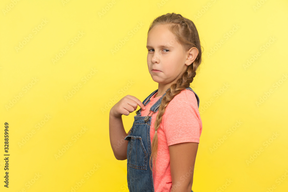 This is me! Portrait of egoistic self-confident little girl in denim overall pointing herself and looking with arrogance, proud of achievement success. indoor studio shot isolated on yellow background This is me! Portrait of egoistic self-confident little girl in denim overall pointing herself and looking with arrogance, proud of achievement success. indoor studio shot isolated on yellow background
