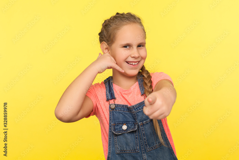 Hey you, let's communicate! Portrait of funny positive little girl with braid in denim overalls making call with fingers shaped like telephone and pointing to camera. indoor studio shot, isolated Hey you, let's communicate! Portrait of funny positive little girl with braid in denim overalls making call with fingers shaped like telephone and pointing to camera. indoor studio shot, isolated