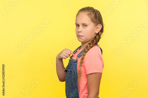This is me! Portrait of egoistic self-confident little girl in denim overall pointing herself and looking with arrogance, proud of achievement success. indoor studio shot isolated on yellow background