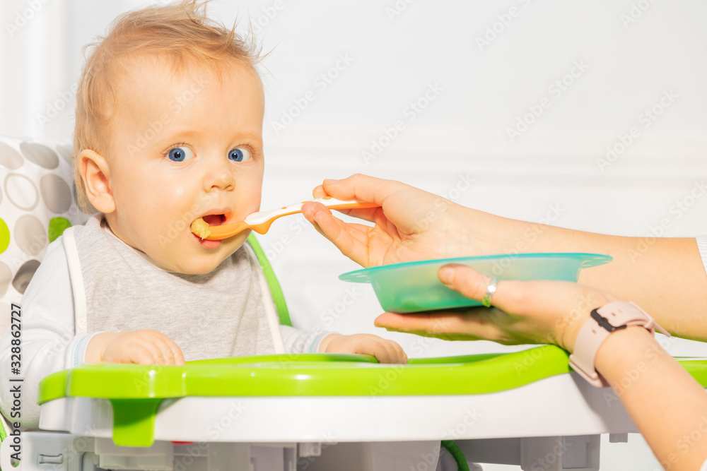 Happy little toddler baby boy sit in high chair and eat from plastic spoon in mouse looking at camera Happy little toddler baby boy sit in high chair and eat from plastic spoon in mouse looking at camera