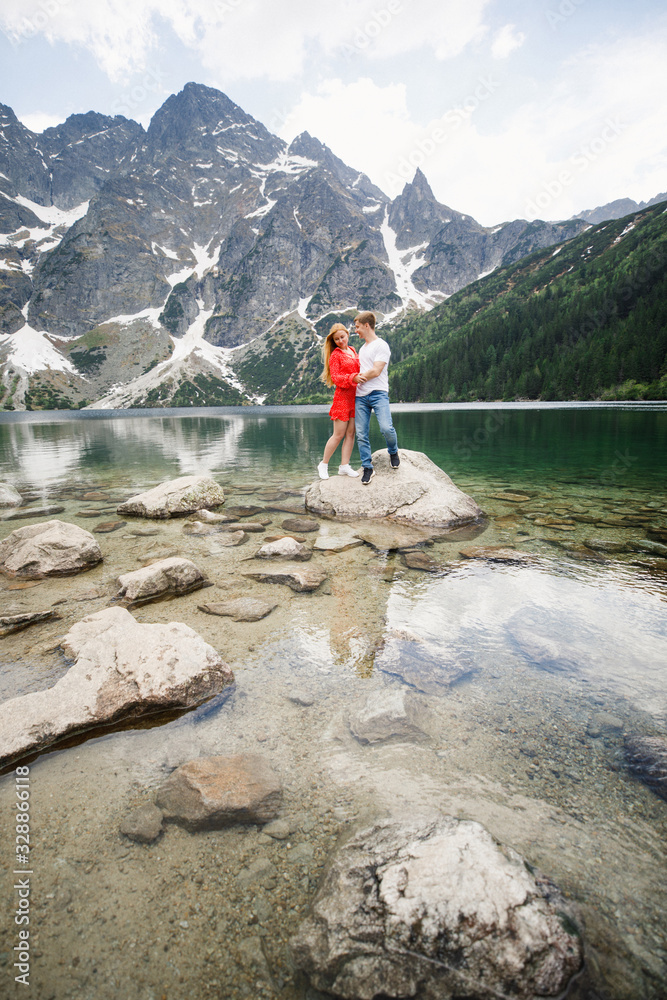 a young couple, a girl in a red dress, a man in a white T-shirt and blue pants stand on a background of mountains near the lake in the park a young couple, a girl in a red dress, a man in a white T-shirt and blue pants stand on a background of mountains near the lake in the park