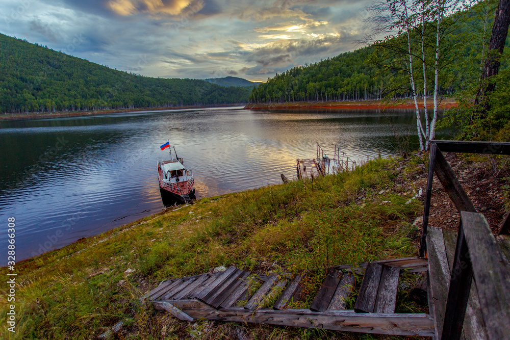 Beautiful landscape. Zeya reservoir, Amur region. A small boat moored at the reserved shore against the backdrop of a beautiful sunset. Reflection of sunset in the water. Beautiful landscape. Zeya reservoir, Amur region. A small boat moored at the reserved shore against the backdrop of a beautiful sunset. Reflection of sunset in the water.