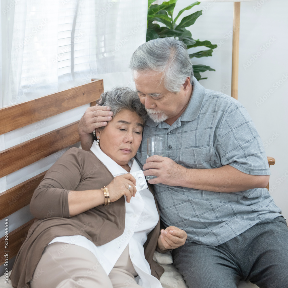 Obraz premium Asian Senior Woman taking medicines and drinking water while sitting on couch. Old man take care his wife while her illness at the house.