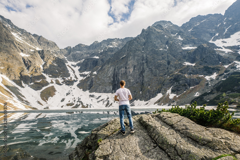 a man in a white T-shirt and blue pants stands on a stone and looks at a lake and mountains in the Tatra National Park in Poland